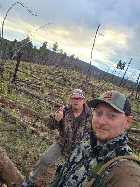 Two men in camouflage and outdoor gear taking a selfie in a forested area with fallen trees and sparse trees, under a cloudy sky.