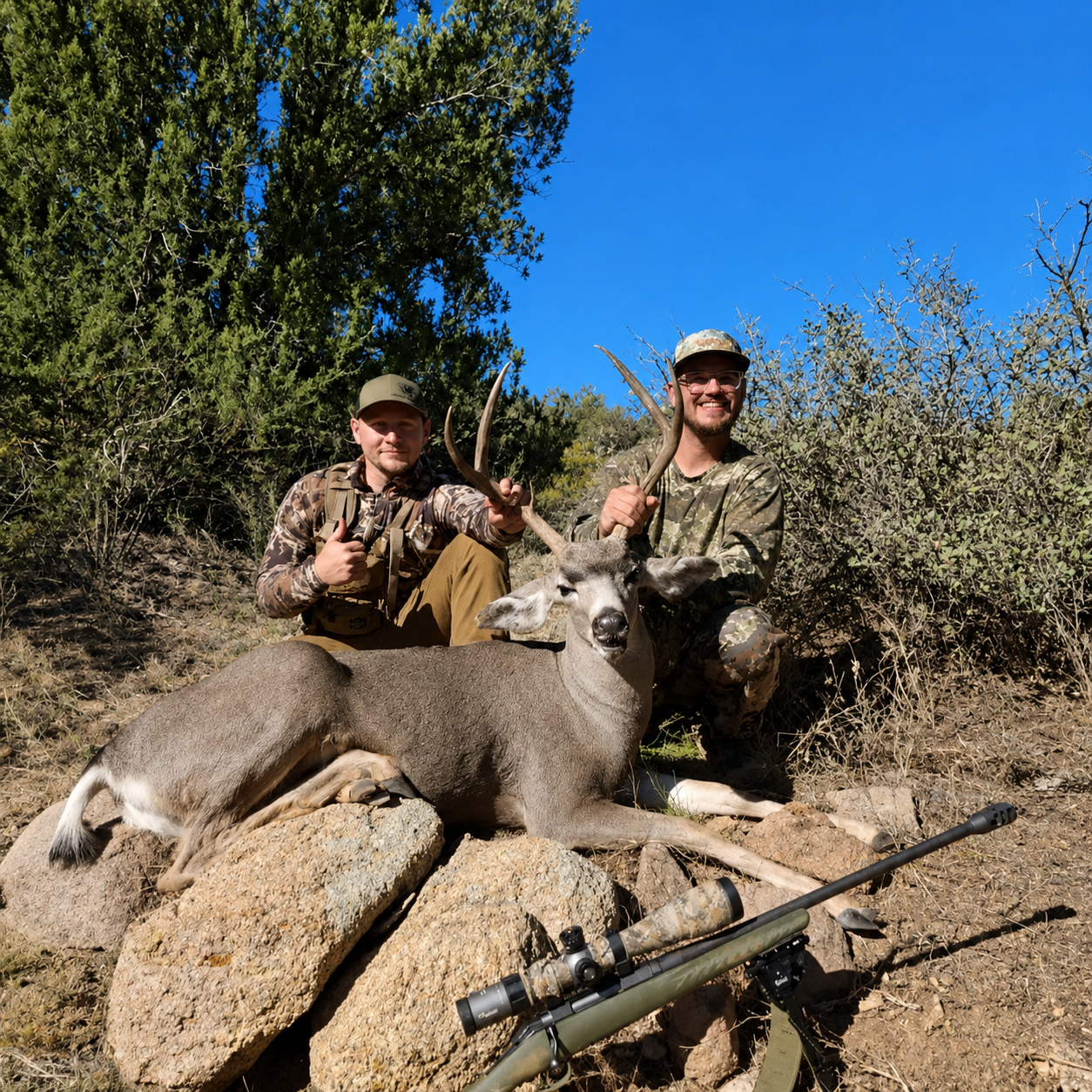 Two hunters kneel on rocks in a desert landscape, each holding the antlers of a deer they have hunted. One man has a beard and glasses, wearing camouflage clothing, and the other is younger, in a camouflage jacket and hat. A rifle with a scope is lyi