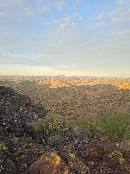 A landscape view of rugged, rocky terrain with sparse desert vegetation under a partly cloudy sky.