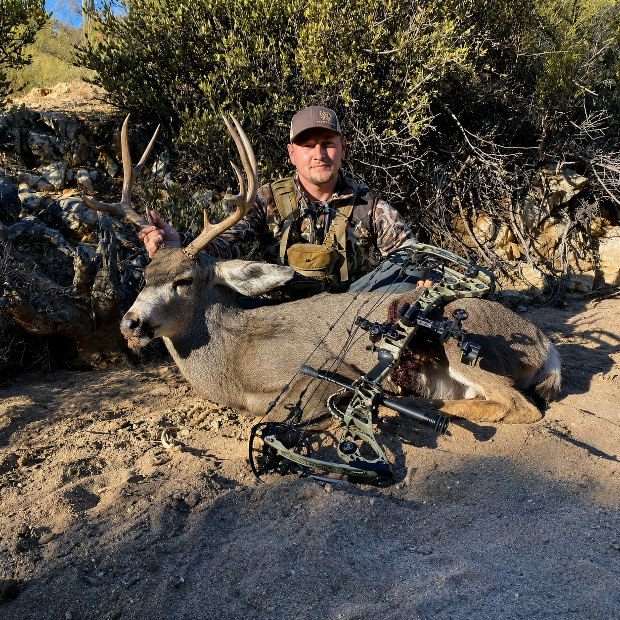 A man in camouflage clothing and a cap sitting on the ground in a desert landscape, holding a large deer with antlers after a hunting trip, with a bow resting on the deer.