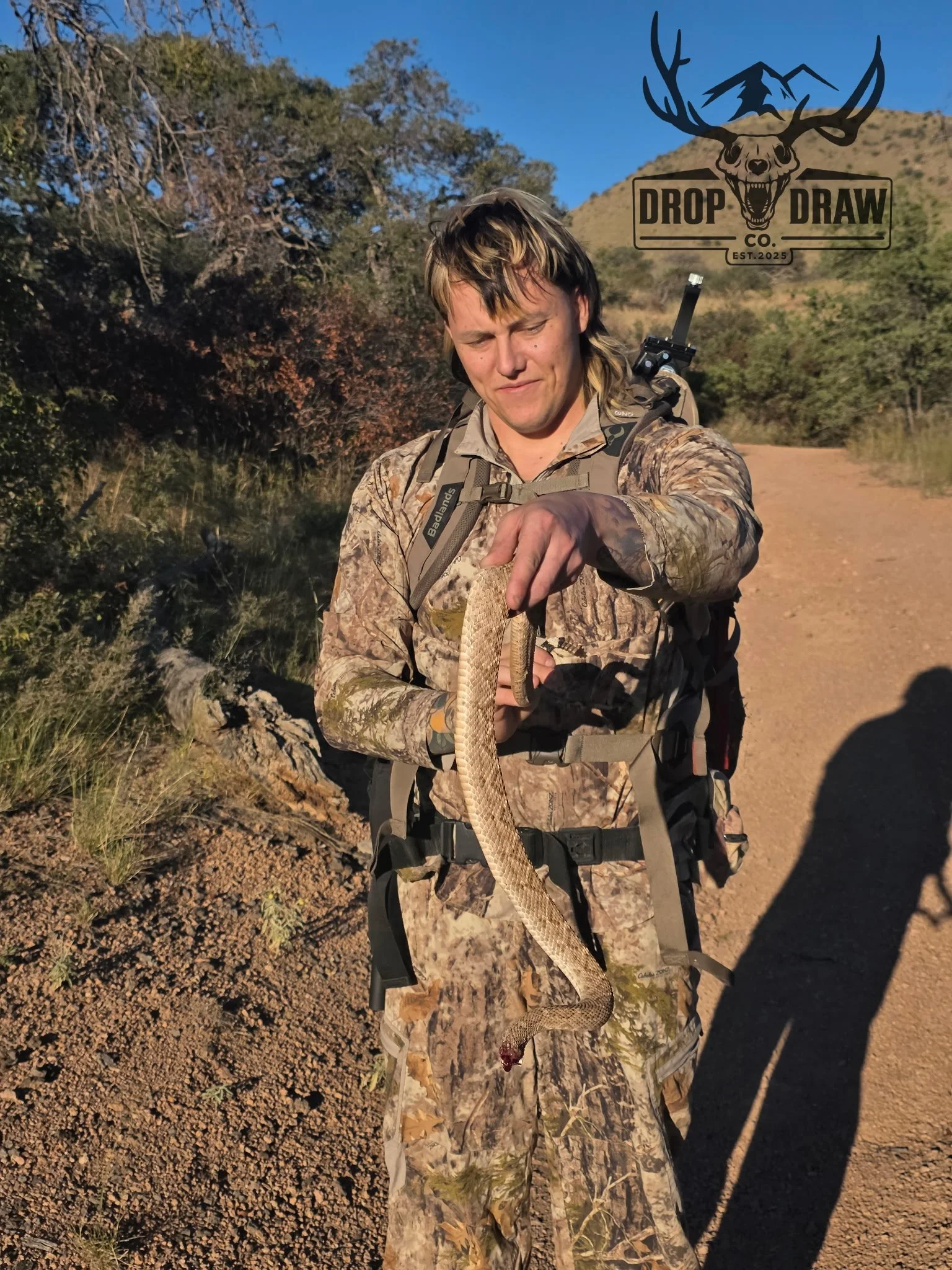 A person in camouflage clothing holding a snake on a dirt trail in a natural outdoor setting with hills and trees in the background.