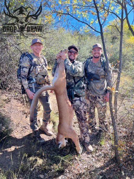 Three men in camouflage and tactical gear standing outdoors, holding a large, freshly hunted animal by its front legs.