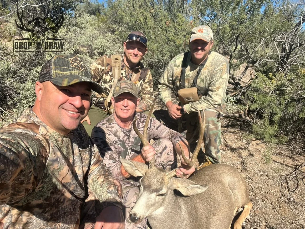Four men in camouflage clothing posing outdoors with a large deer they have hunted, in a dry, bushy area.