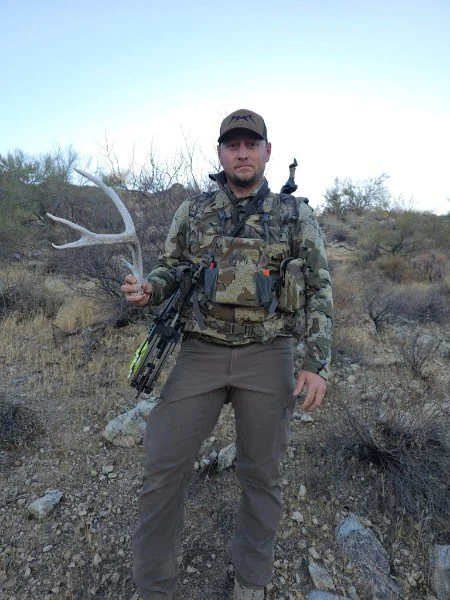Man in camouflage and outdoor gear standing in a desert landscape holding a large antler and a rifle.