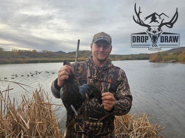 Man in camouflage clothing holding a duck he caught by the water with reeds in the foreground and a cloudy sky, logo of Drop & Draw Outfitters in the top right corner.