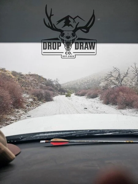 View from inside a vehicle on a dirt road surrounded by bushes and trees, with a logo of a deer skull with antlers and mountains in the background, and the text "Drop Draw Co. EST. 2023"