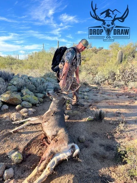 A man with a backpack and cap standing in a desert landscape, holding a hunting rifle with a dead deer on the ground in front of him. The sky is partly cloudy and there are cacti and bushes in the background. A logo in the top right corner features a