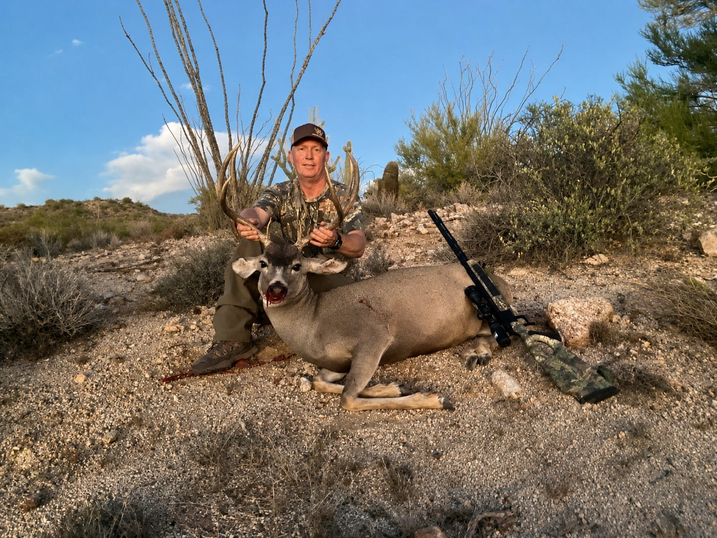 A man in camouflage clothing holding the antlers of a large deer he has hunted and lying on the desert ground in a dry, shrub-filled landscape. There is a rifle with a scope resting on the ground next to the deer.