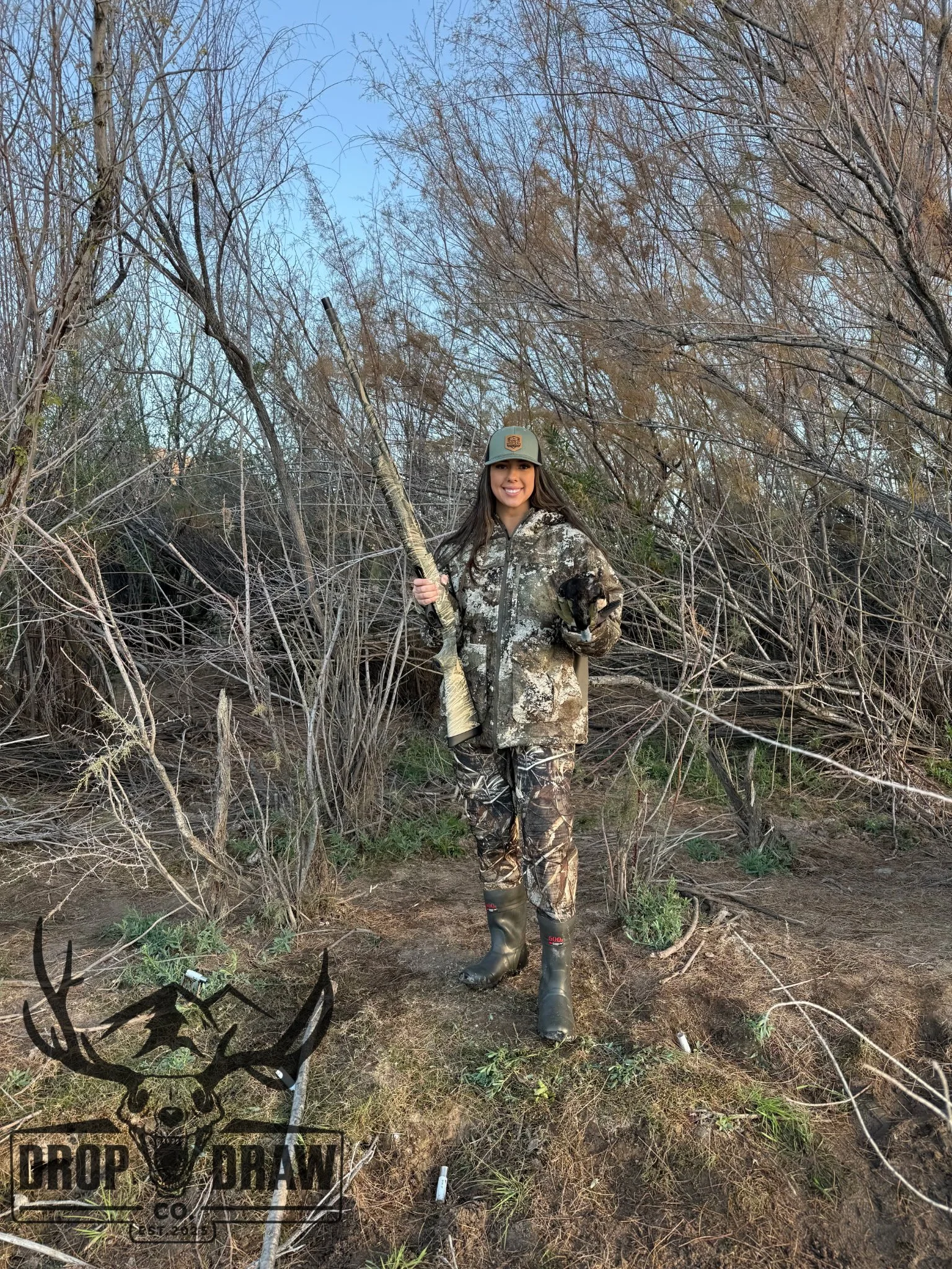 Woman in camouflage jacket and boots holding a rifle in a wooded area with leafless trees and a blue sky.