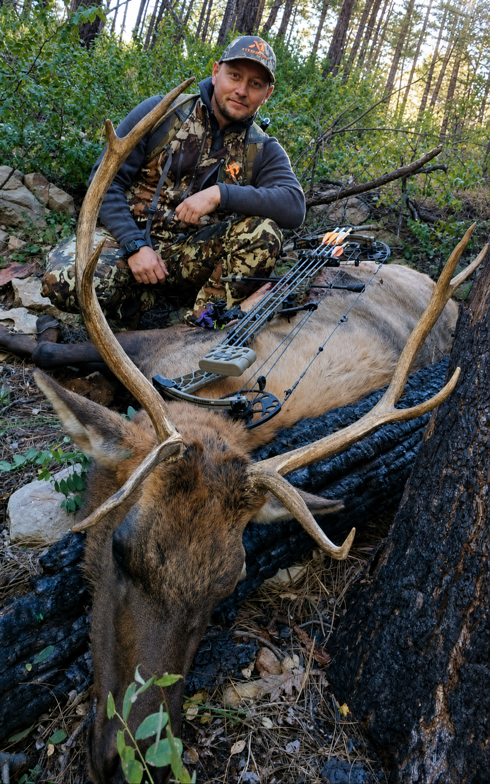 A man in camouflage clothing and a cap is kneeling beside a large deer with impressive antlers, which has been hunted and laid on the ground in a forest. The man has a bow and arrow resting on the deer's body.