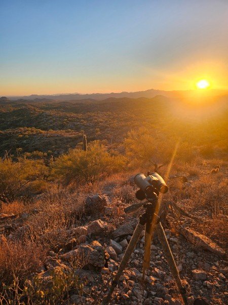 A telescope set up on a tripod overlooking a desert landscape at sunset with hills and cacti.