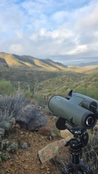 A pair of binoculars mounted on a tripod observing a mountainous landscape with hills and a partly cloudy sky.