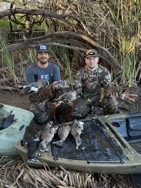 Two men in camouflage clothing kneeling in a marshy area with tall grass and branches, holding ducks and hunting supplies near a kayak. Six hunted ducks and two raccoons are displayed on the kayak.