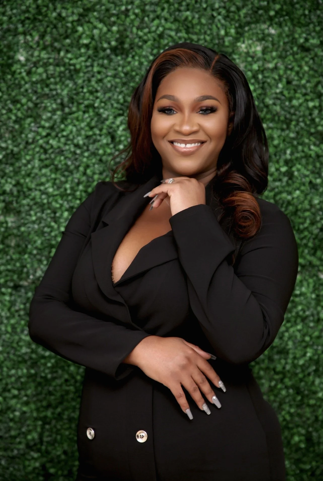A woman with long, dark hair and a warm smile, wearing a black blazer and standing in front of a green, leafy background.
