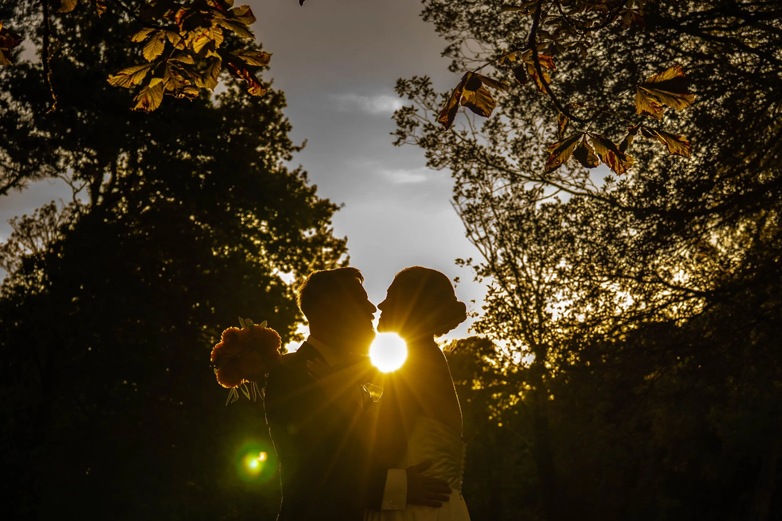 A couple is kissing outdoors during sunset, framed by trees and leaves, with the sun shining behind them creating a silhouette effect.