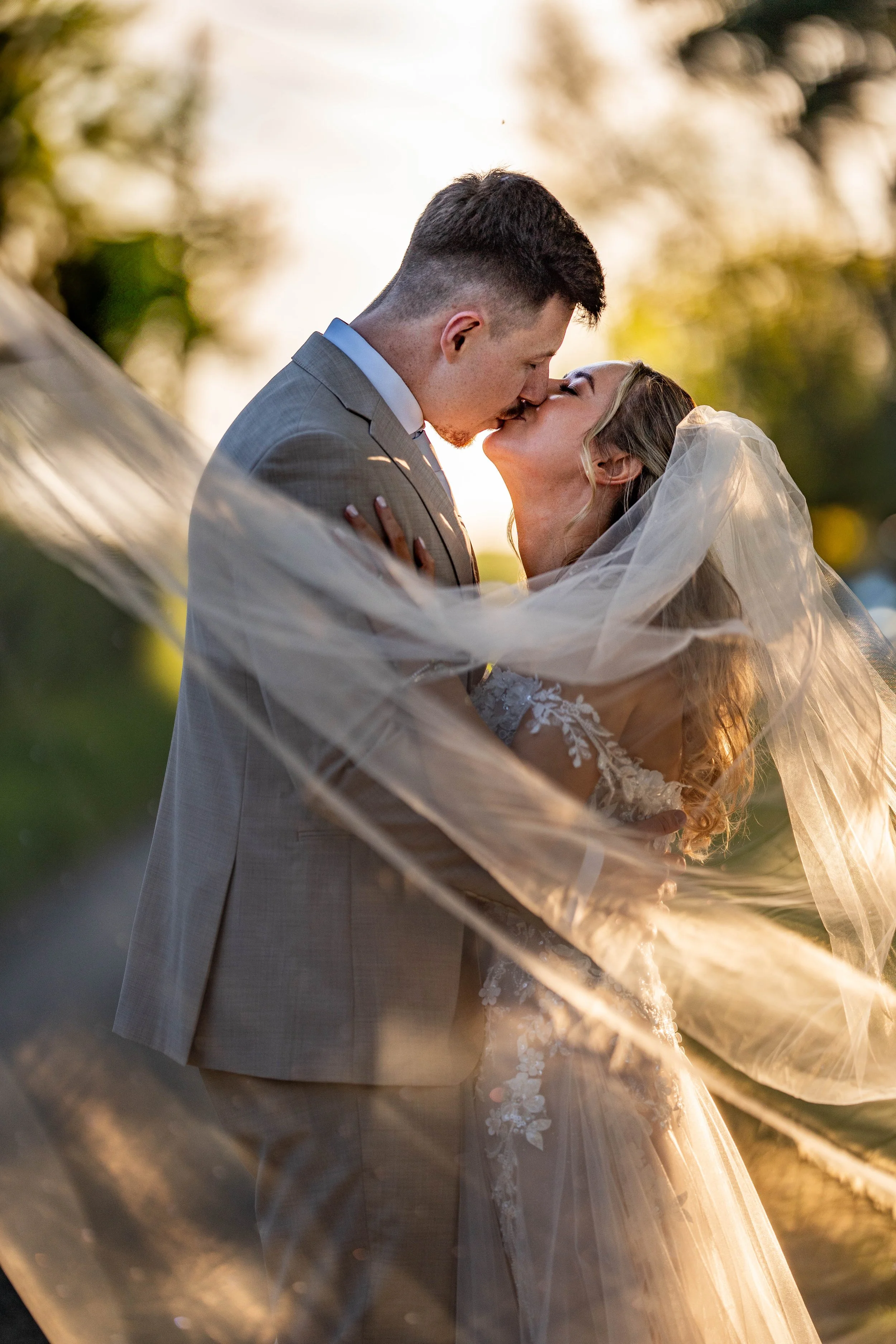 A couple in wedding attire sharing a kiss outdoors during sunset, with blurred trees and sunlight in the background.