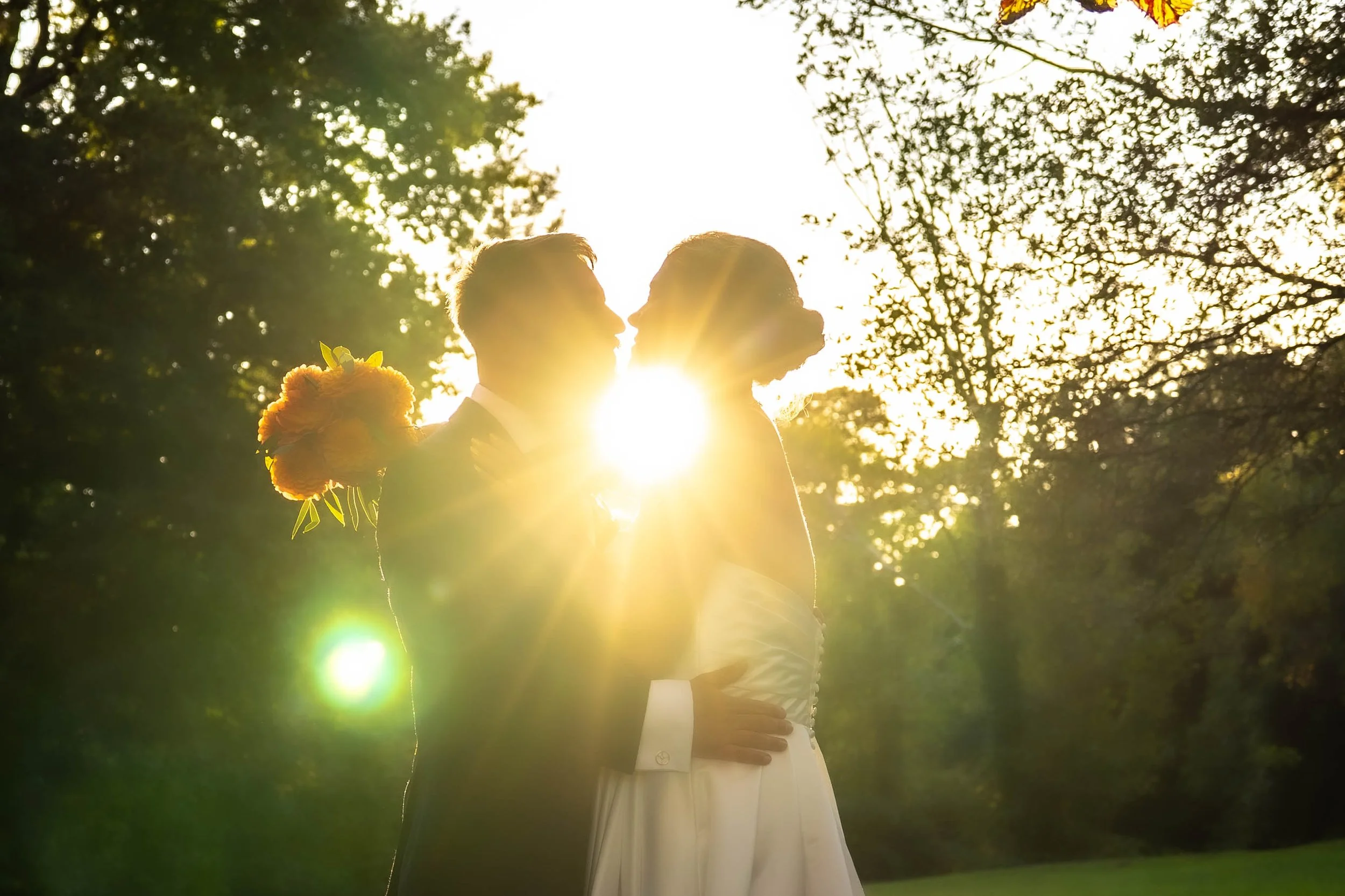 A couple in wedding attire facing each other and touching foreheads outdoors with trees in the background, backlit by the setting sun.