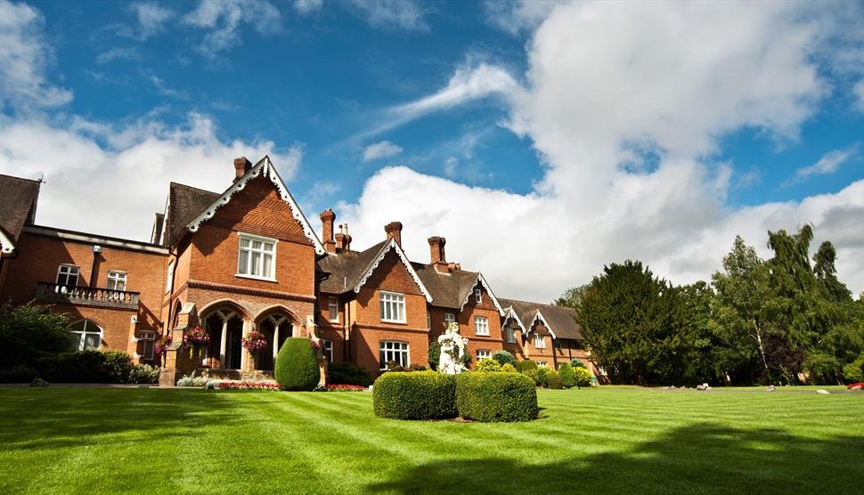 A large brick manor house with a manicured lawn, lush trees, and a partly cloudy sky.