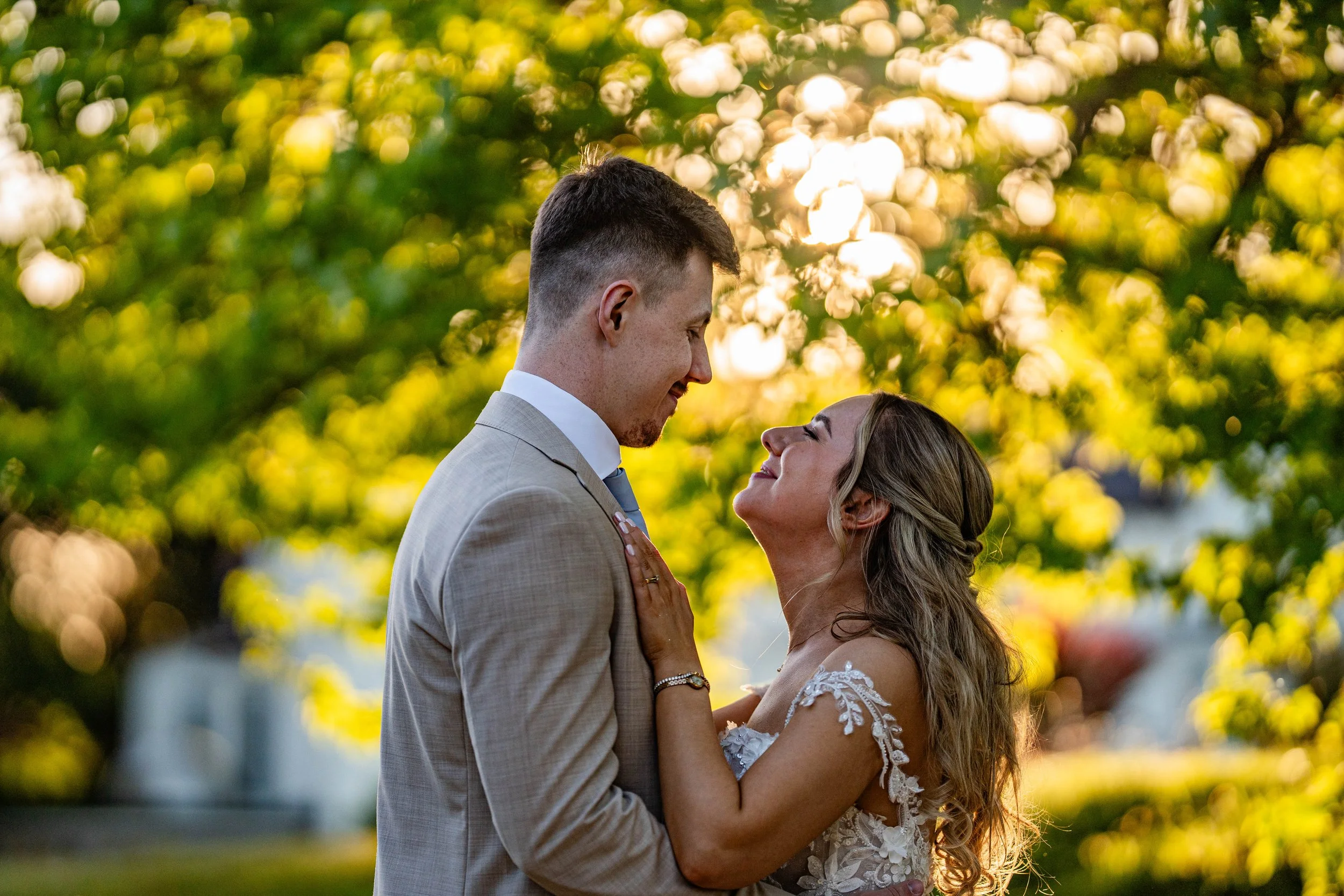 A groom in a beige suit and a bride in a lace wedding dress holding each other and smiling in an outdoor setting with a blurred background of trees and sunlight.