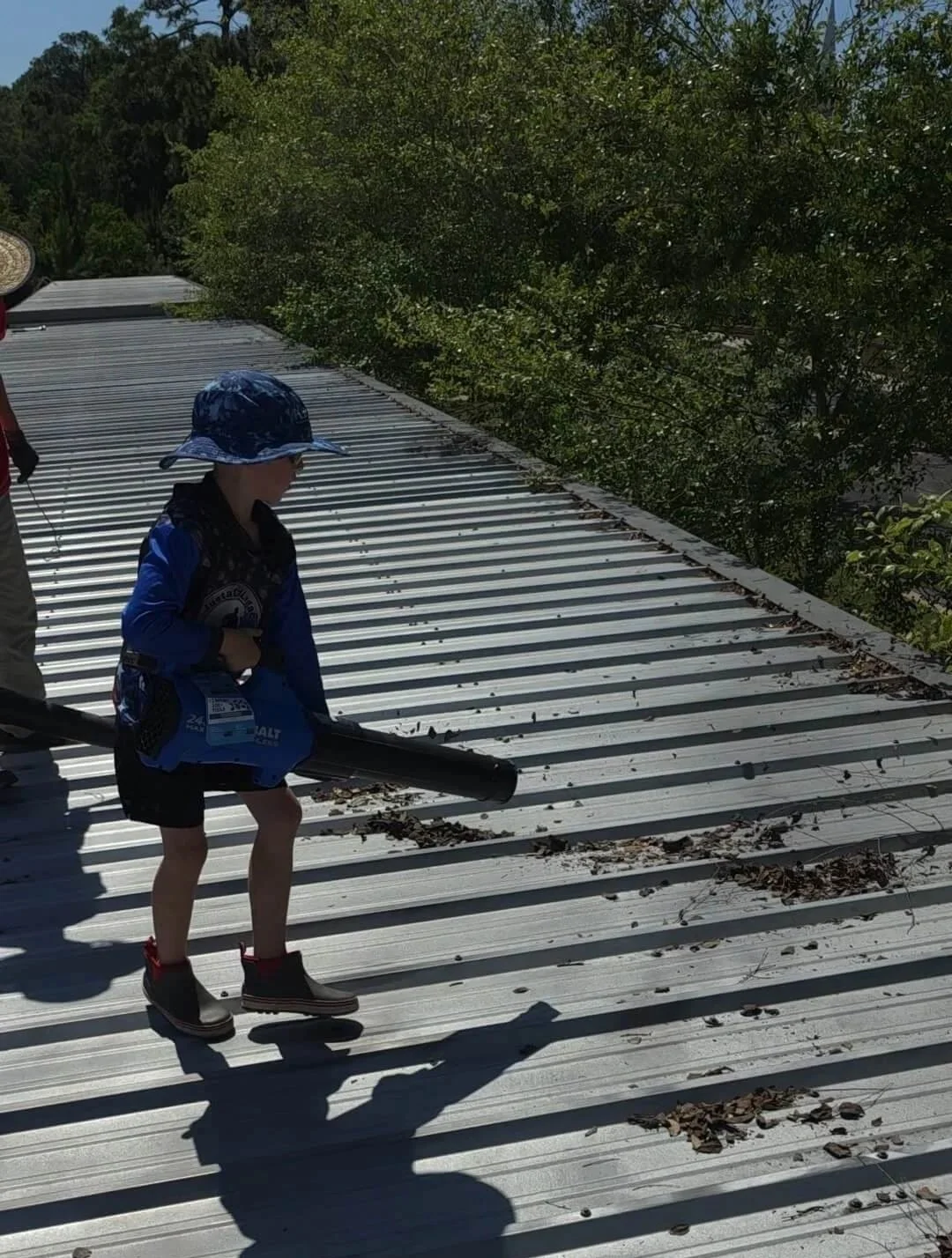 Child wearing hat and boots using leaf blower on metal roof, with trees in background.