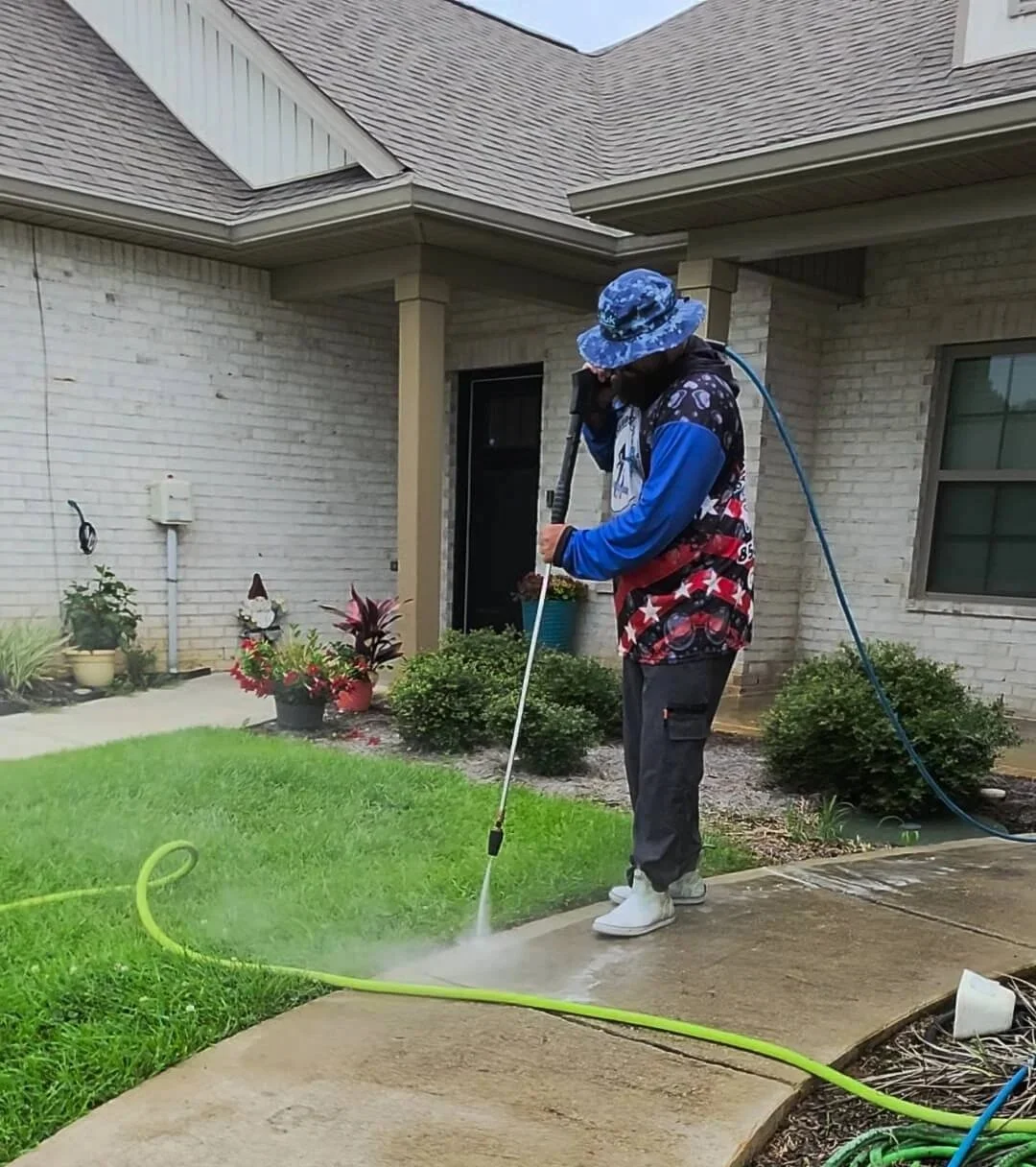 Person pressure washing sidewalk in front of house, wearing hat and casual clothes.