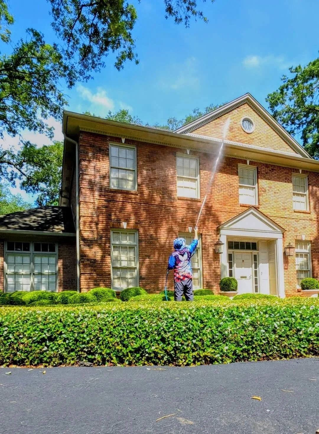 A person standing on a neatly manicured lawn, watering the side of a large brick house under a blue sky.