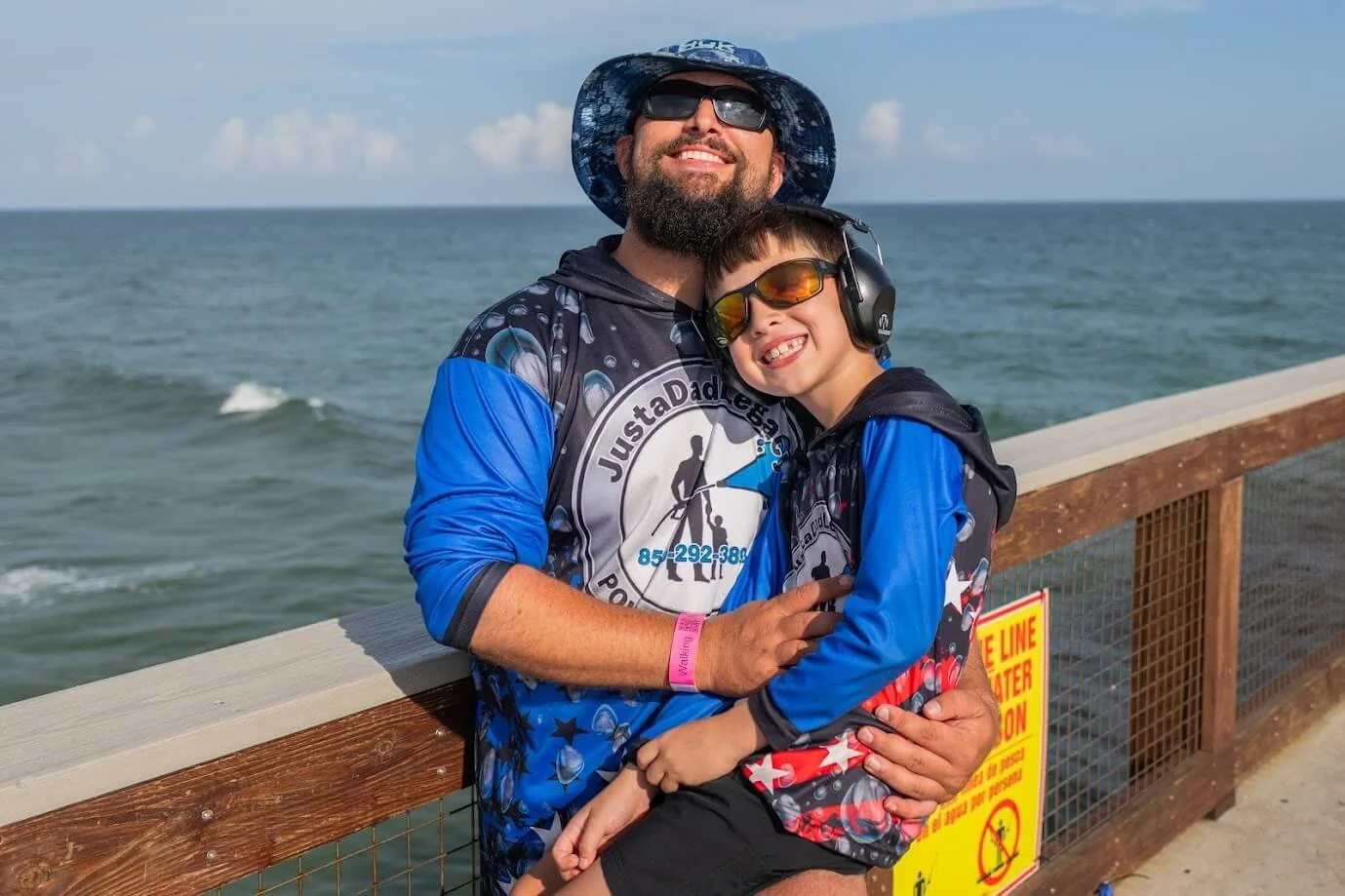 A man and a young boy smiling and hugging on a pier near the ocean. The man is wearing sunglasses, a sun hat, and a shirt with water and logo designs. The boy is wearing sunglasses, headphones, and a blue hoodie with a patriotic design, with an American flag pattern. There is a yellow warning sign on the pier and an ocean with waves in the background.
