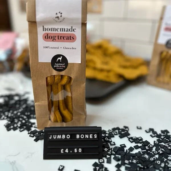 A package of homemade dog treats labeled as 100% natural and gluten-free, with a small window showing the treats inside, placed on a surface with scattered black letter tiles and a black board displaying "JUMBO BONES" and the price "£4.50".