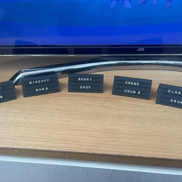Small black letter boards with words such as "BISCOFF BOMB," "BERRY DROP," "CREME CRUMB B," and "CLASS C RUSH" on a wooden surface in front of a blue monitor.