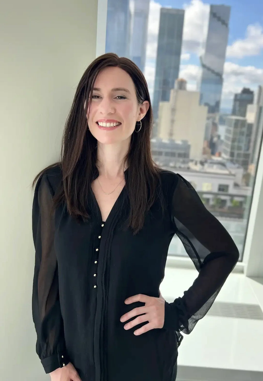 A woman with long dark hair wearing a black blouse with sheer sleeves and gold button details, smiling with a city skyline view in the background.