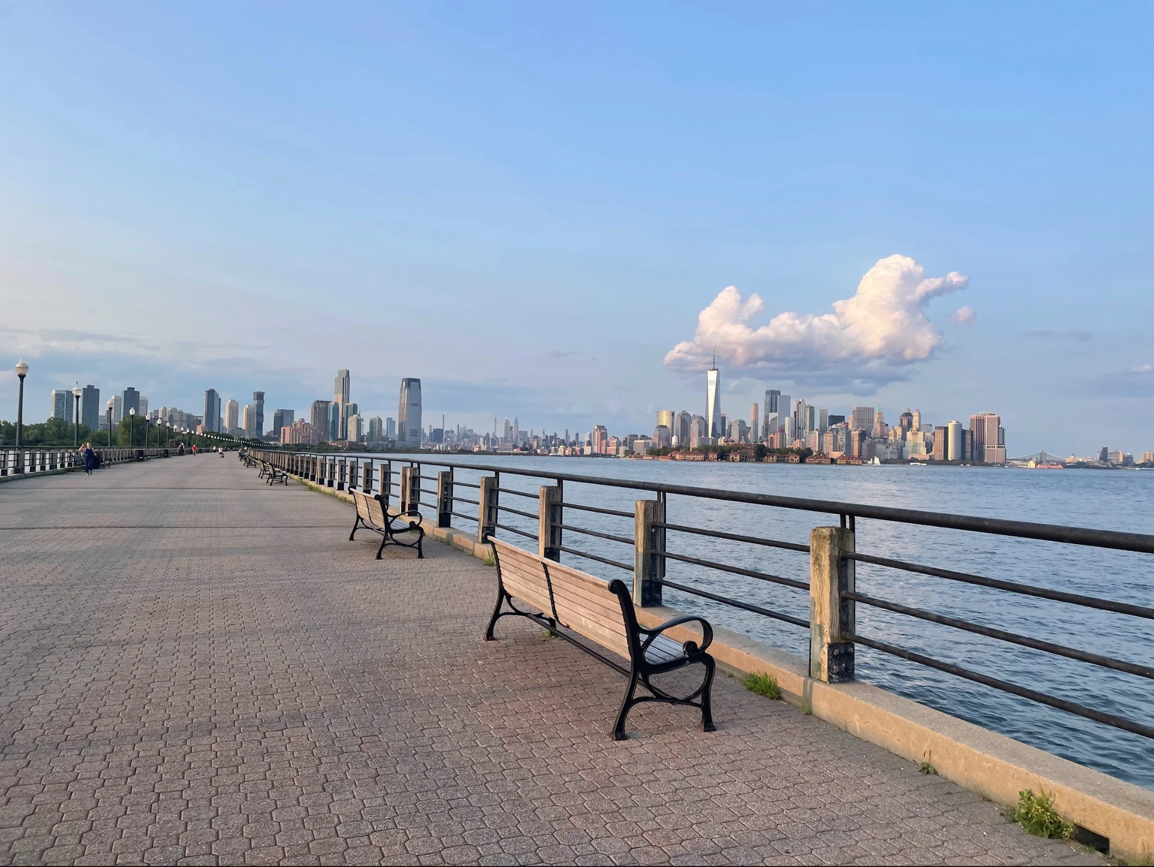 A waterfront promenade on a clear day, with benches and a metal railing along the edge, overlooking a city skyline with tall buildings and a body of water.