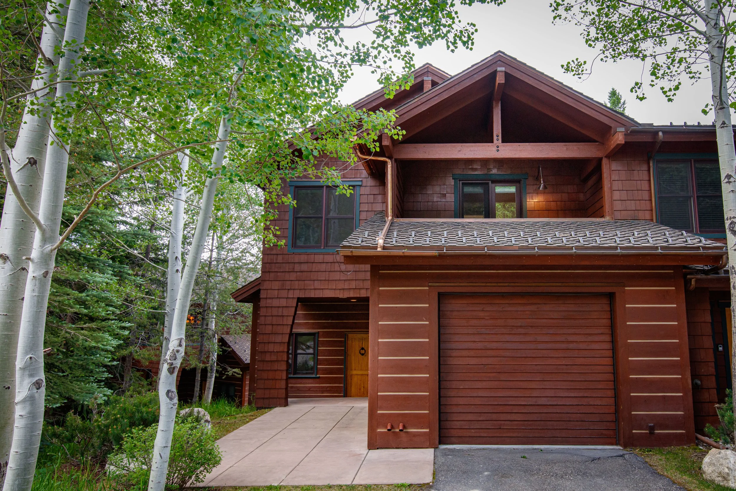 The image shows the front of a two-story house with a wooden exterior, surrounded by tall, leafy trees. The house has a sloped roof with visible wooden beams and a small covered porch area at the entrance. There is a paved walkway leading to the front door, and the house is nestled in a lush, green natural setting.