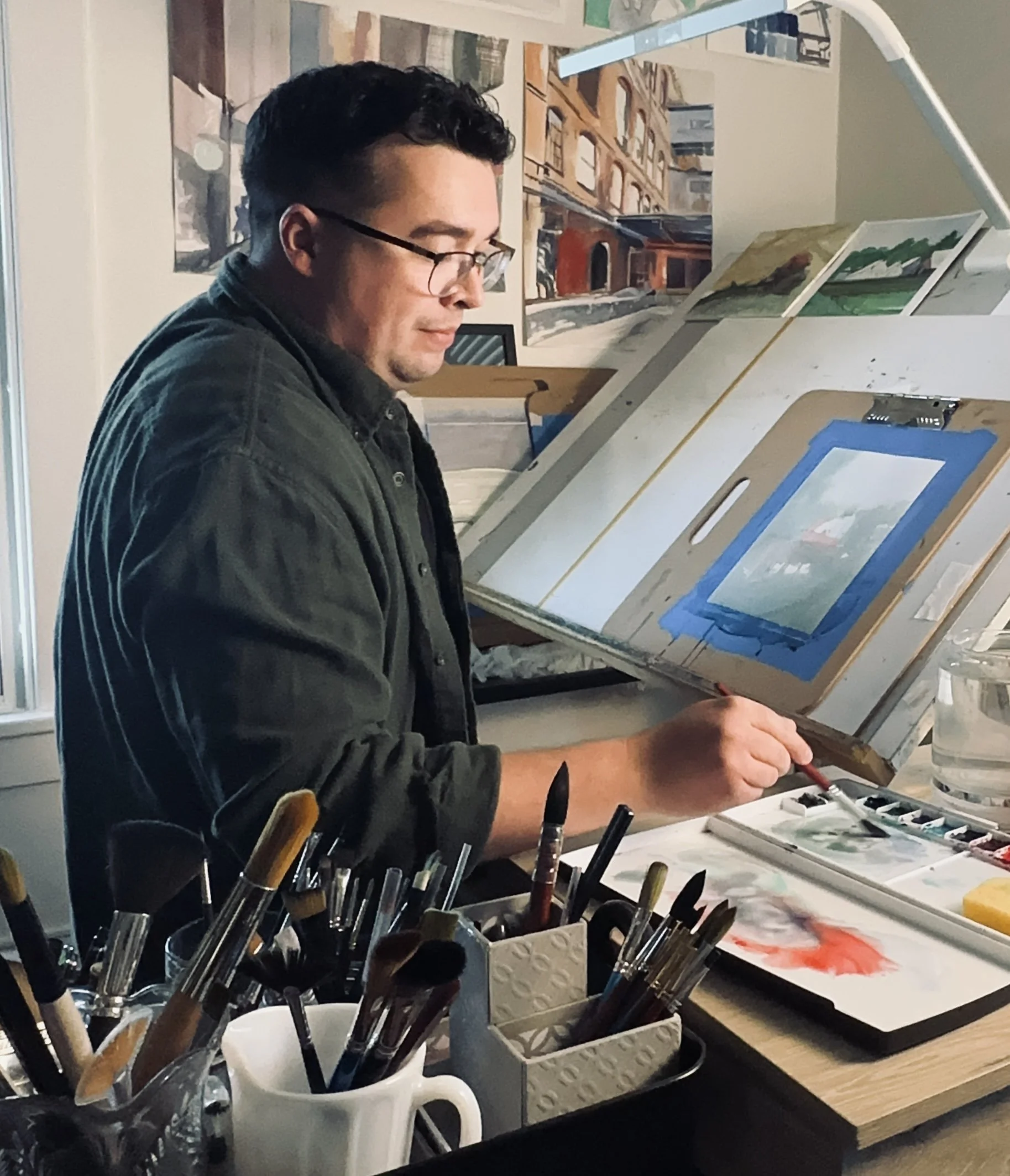 An artist in a studio, painting detailed urban scenes on a white canvas. Art supplies, including brushes and watercolors, are organized on a table nearby, with artwork displayed on the wall in the background.
