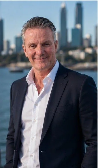A middle-aged man in a white shirt and dark blazer smiling, with a city skyline in the background.