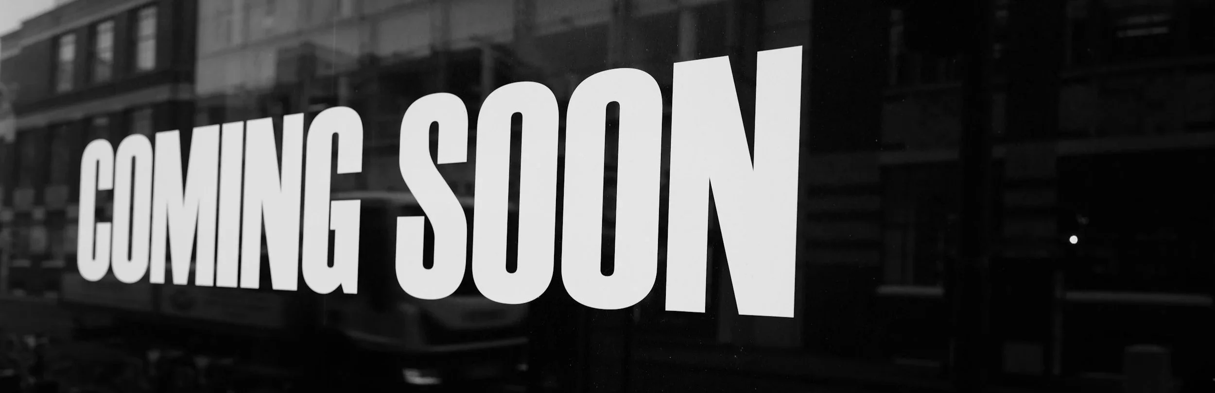 Close-up of a window with the words 'COMING SOON' in large white letters, with reflected buildings in the background.