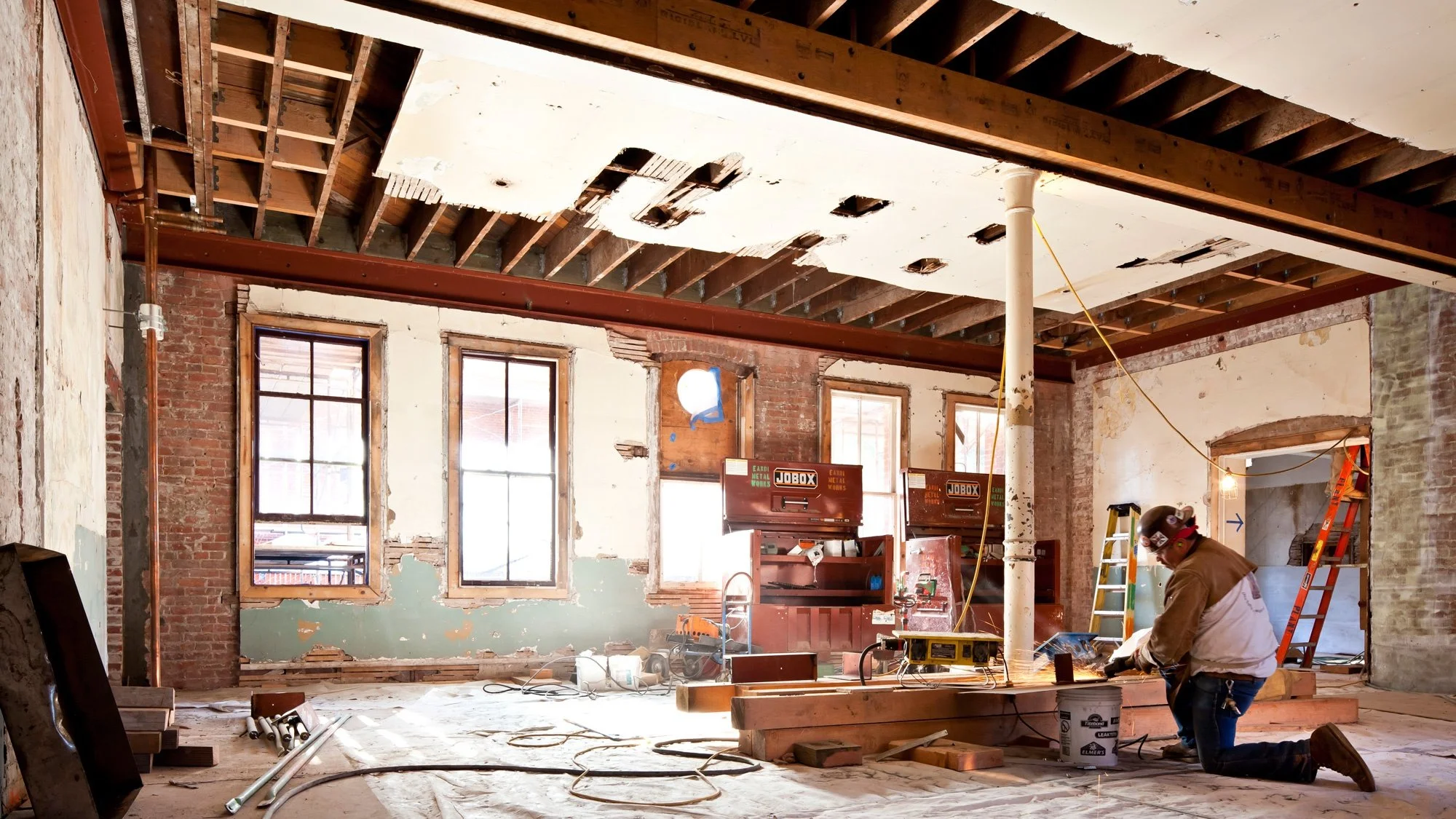 Interior of a building under renovation with exposed brick walls, open ceiling framing, construction tools, ladders, and a worker installing or repairing a wall.