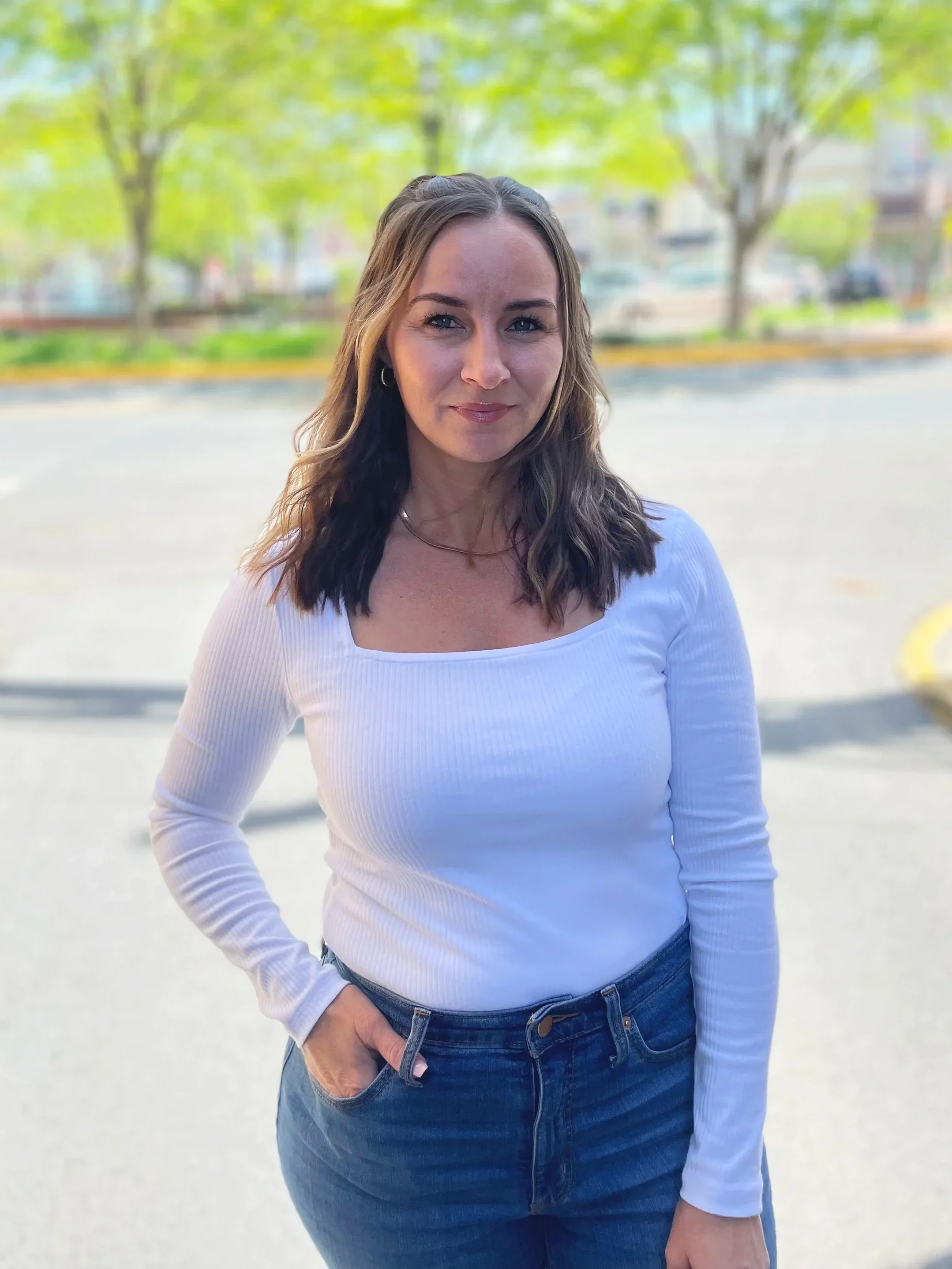 A woman with wavy brown hair and blue eyes wearing a white long sleeve shirt and jeans, standing outdoors with green trees and cars in the background.