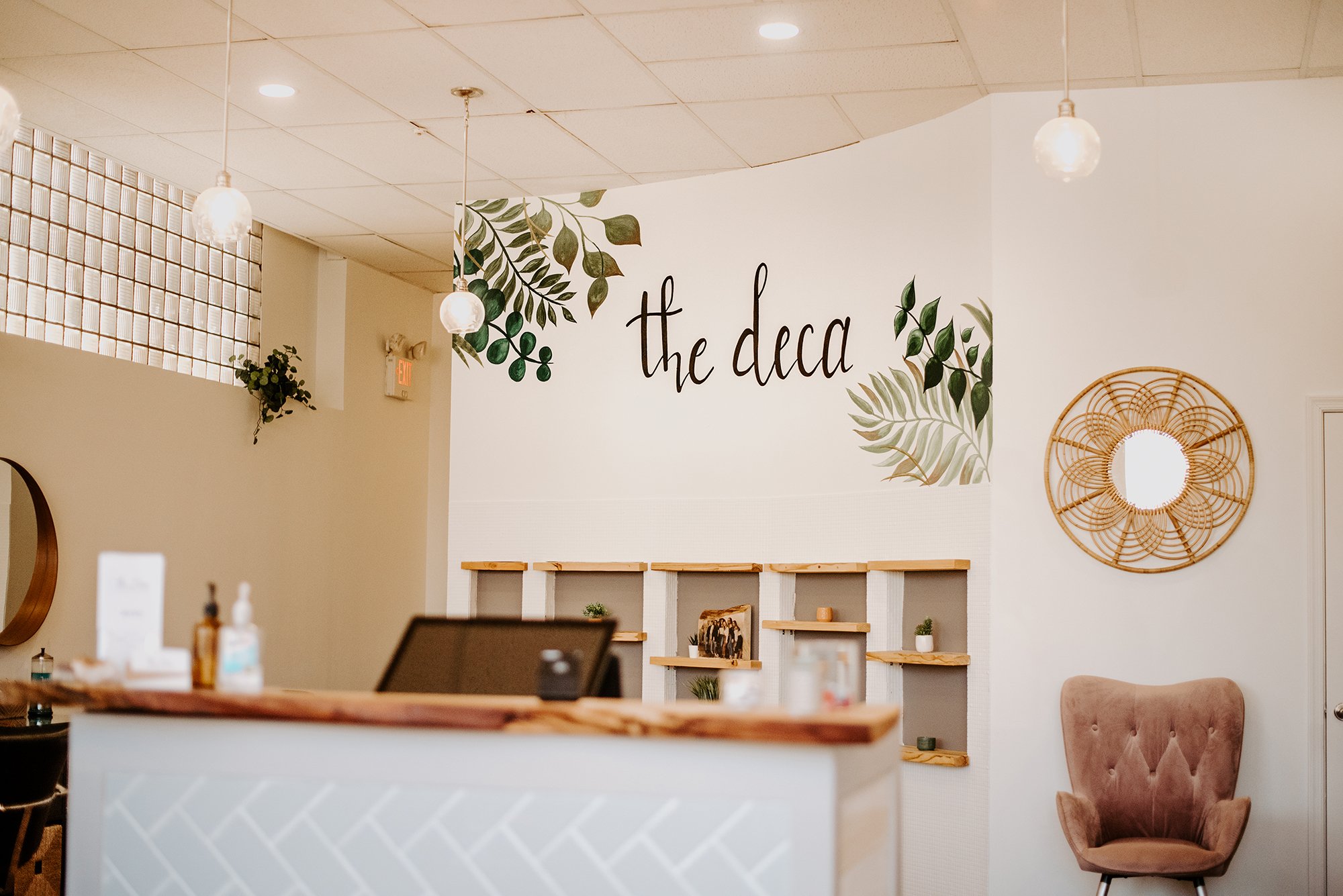Interior of a cozy cafe or boutique, with a large white wall decorated with green leafy accents and the words 'the deca' written in black cursive. There are hanging pendant lights, potted plants, a rattan wall decoration, and a cushioned chair visible in the space.