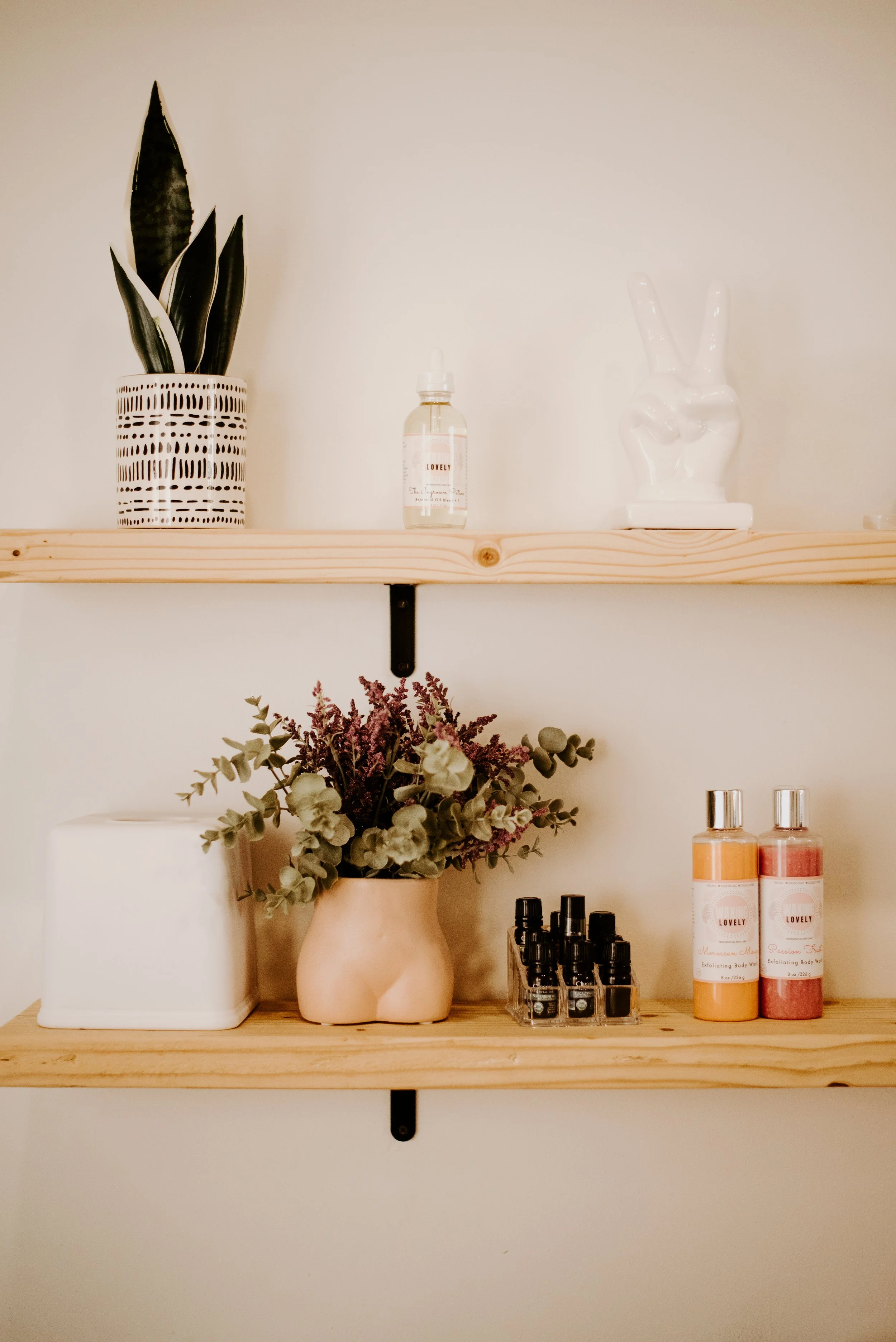 A wooden shelf holding a potted snake plant in a white decorative pot, a small bottle with a dropper, a white ceramic hand sculpture making a peace sign, a vase with green and purple dried flowers, and several bottles of skincare or body products on a lower surface.