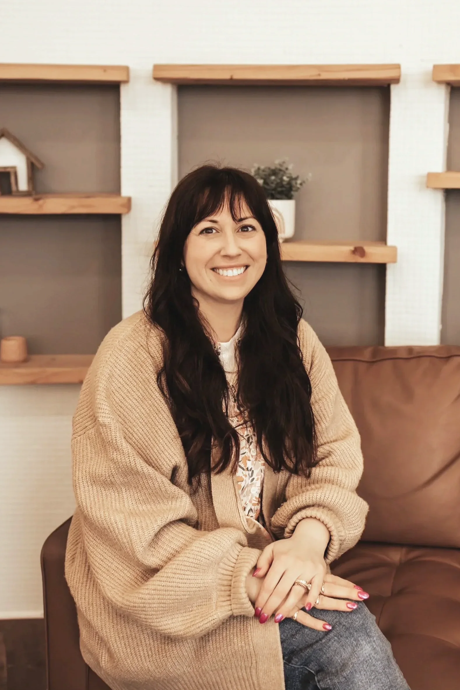 A woman with long dark hair and a beige cardigan sitting on a brown couch, smiling at the camera with hands resting on her lap, in front of a wall with wooden shelves and a small potted plant.