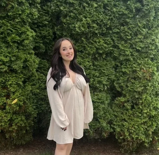 A young woman with long dark hair, smiling, standing in front of green bushes, wearing a light-colored dress.