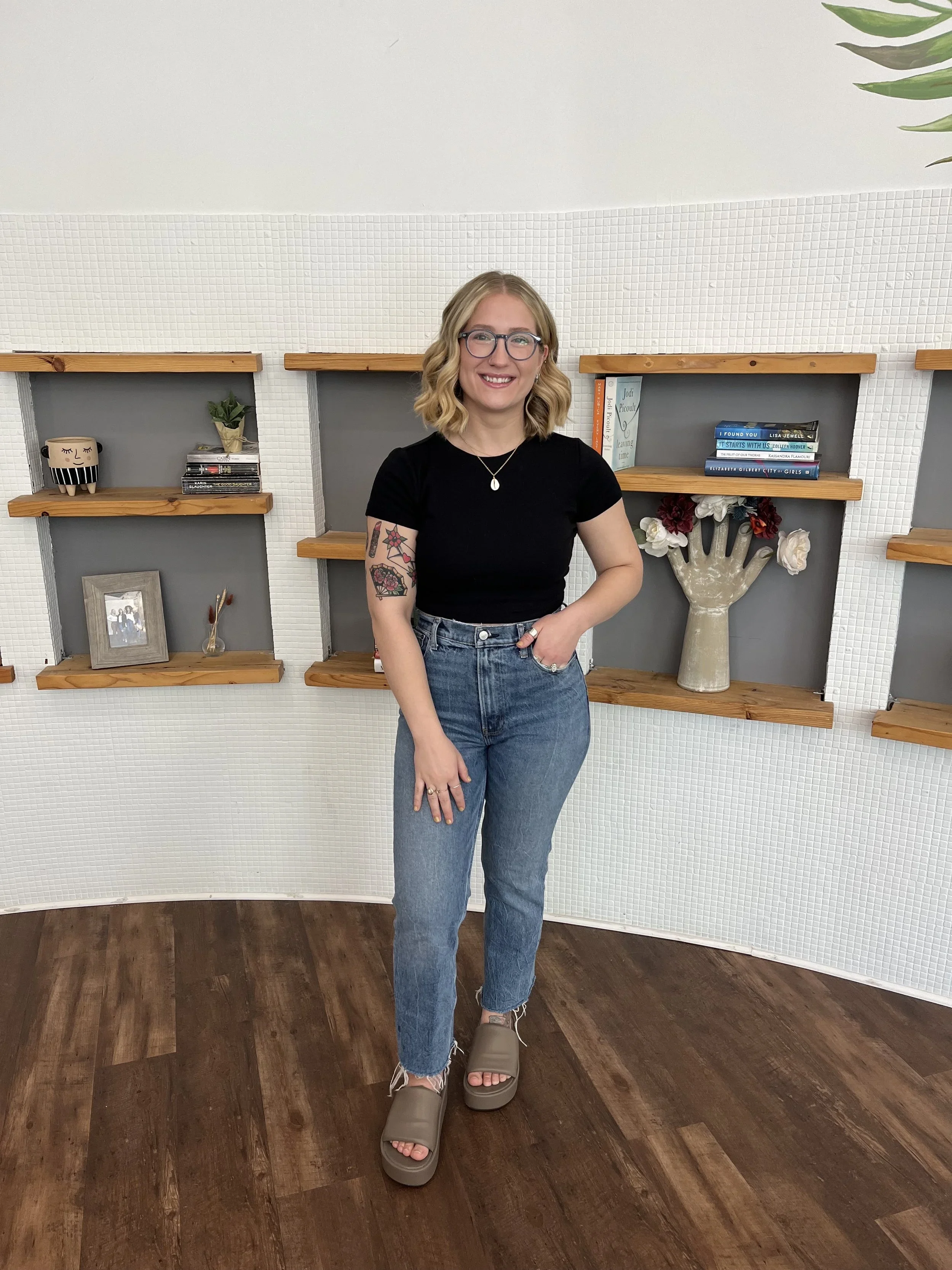 A young woman with glasses and wavy blonde hair stands in front of white textured wall shelves decorated with books, a vase with flowers, and a framed photo. She wears a black t-shirt, high-waisted jeans, and beige slide sandals.