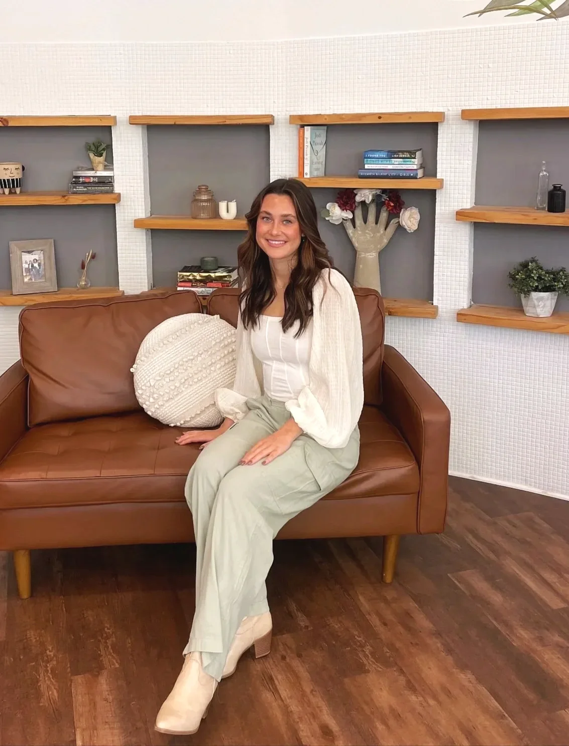 A woman sitting on a brown leather couch with a white round pillow, in a room with wooden shelves and decorative items against a white wall. She has long dark hair, is smiling, and is dressed in a cream-colored blouse and light green pants.