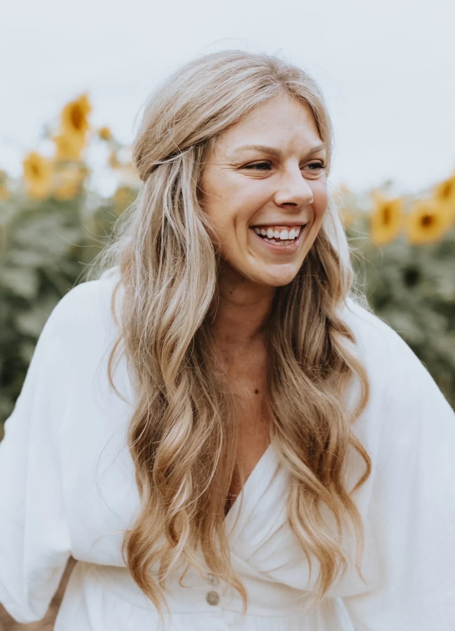 Close-up of a woman with long, wavy blonde hair, smiling outdoors with yellow flowers in the background.