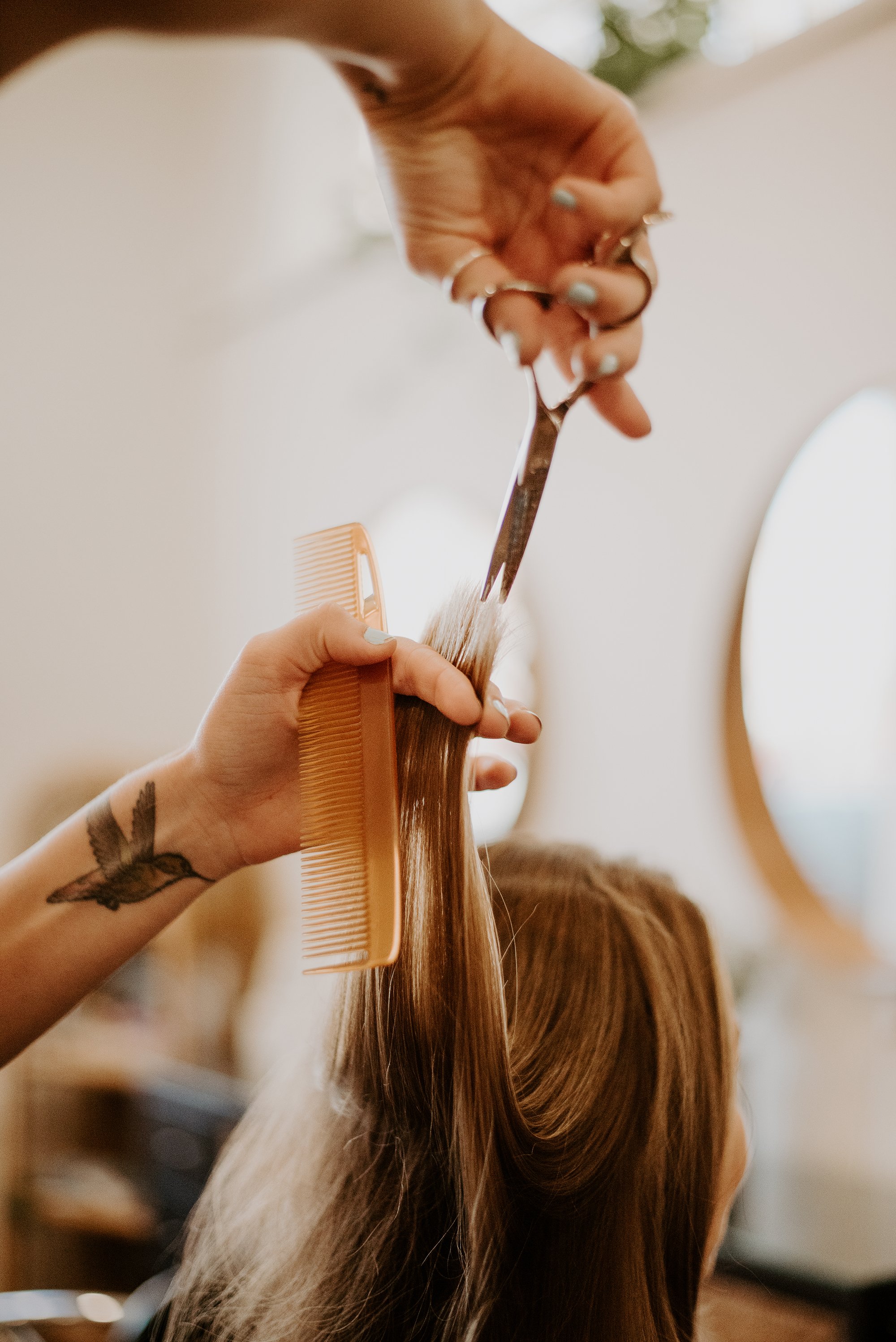 A person with tattoos on their wrist is combing long, straight, reddish-brown hair with a wide-tooth comb and holding scissors in the other hand.