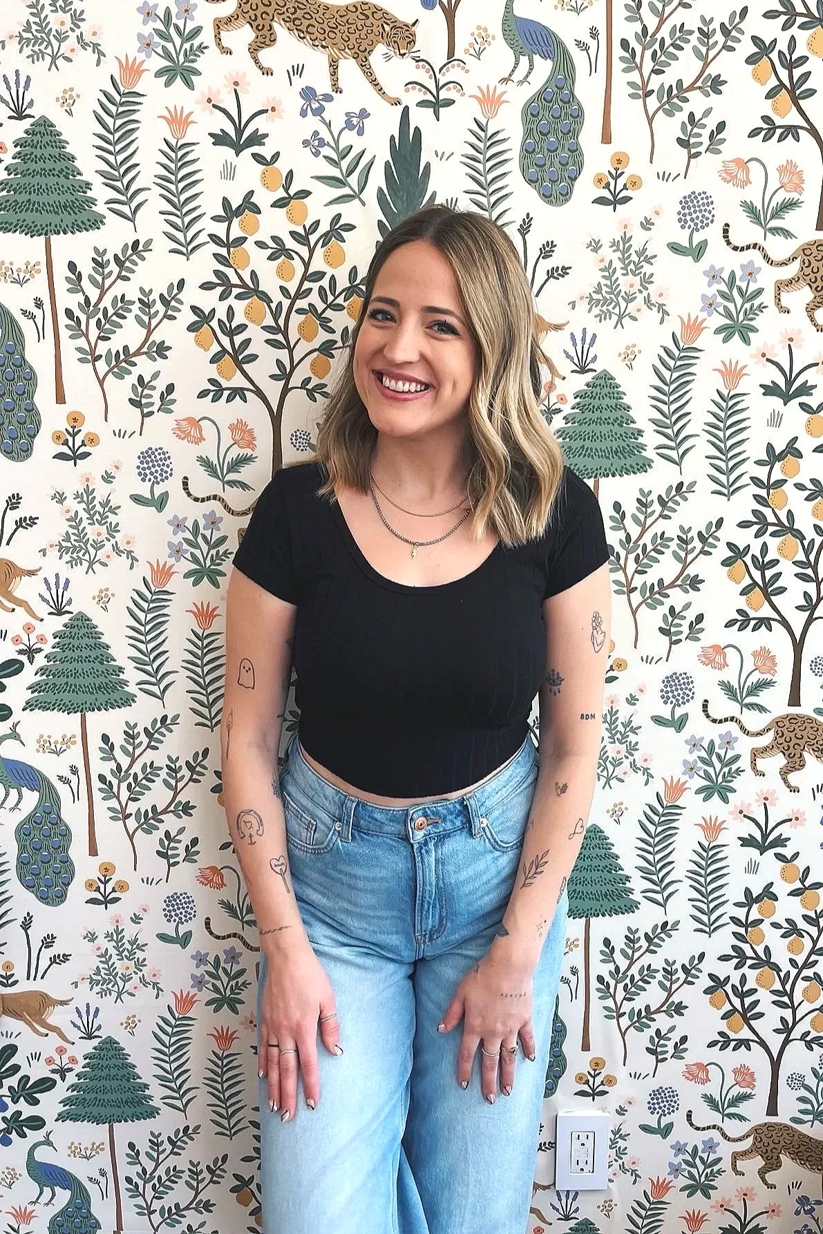 A young woman with blonde hair, smiling, wearing a black t-shirt and light blue jeans, standing in front of a colorful patterned wallpaper featuring jungle animals and plants.