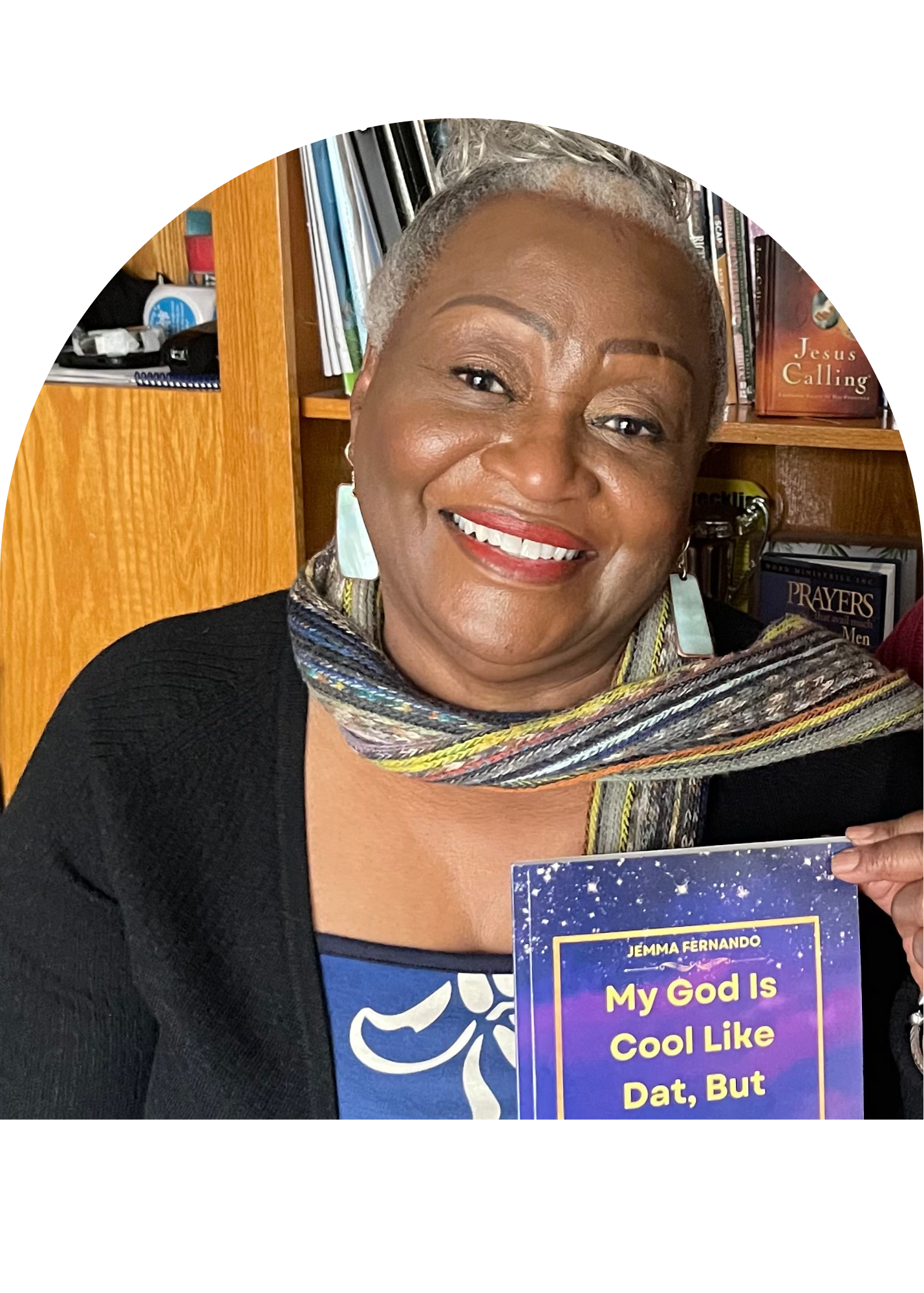 A smiling woman with short gray hair, wearing a colorful scarf and earrings, holding a book titled 'My God Is Cool Like Dat, But' by Jemma Fernando. She stands in front of a bookshelf filled with books, including titles like 'Jesus Calling' and 'Prayers'.