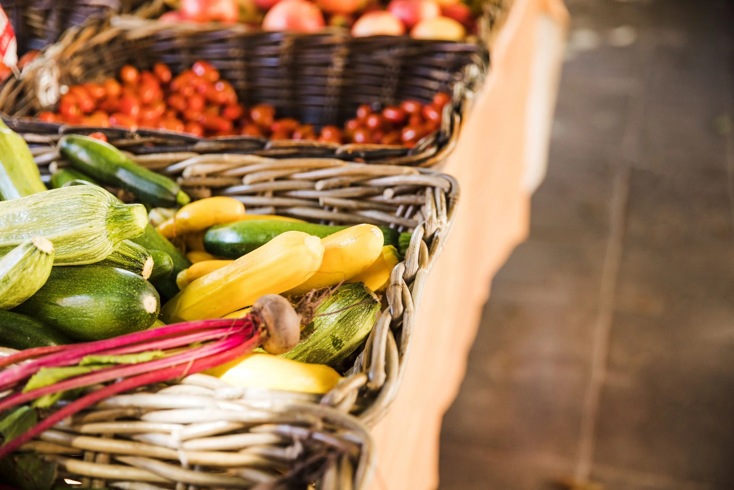A display of fresh vegetables in wicker baskets, including zucchinis, yellow squash, and grape tomatoes.