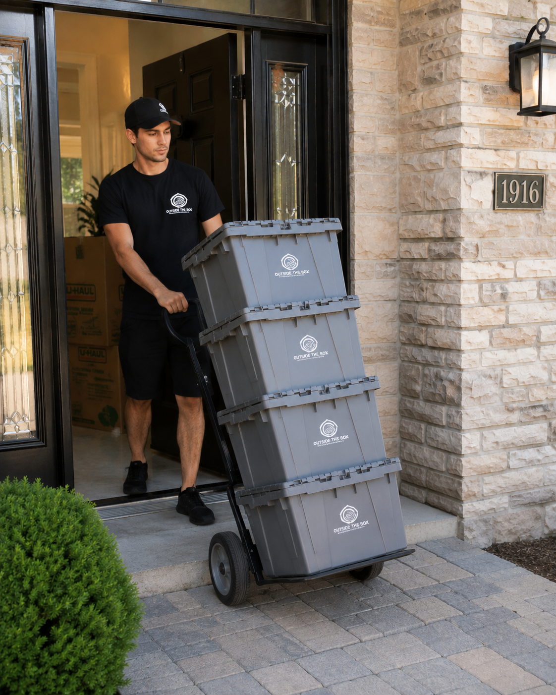 A delivery person is unloading gray trash or recycling bins from a dolly outside a house with stone walls, a black front door, and house number 1916. The bins are labeled 'OUTSIDE THE BOX'.