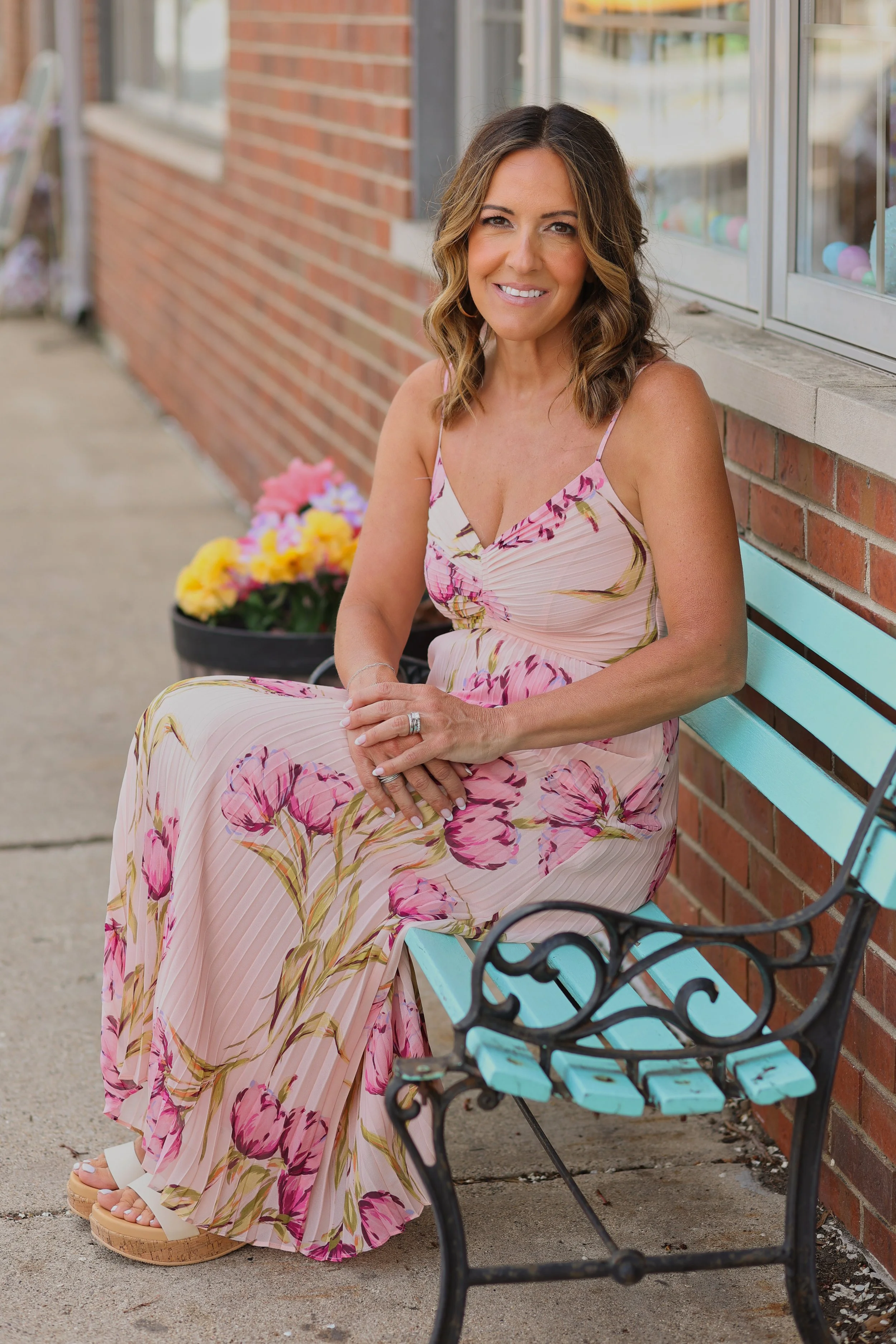 Maria Bullion in a pink floral maxi dress with spaghetti straps sitting on a turquoise park bench outside a brick building, smiling at the camera.